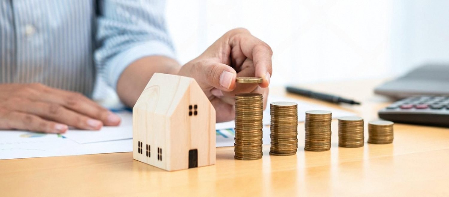 Hand placing coins next to a wooden house model, illustrating home insurance costs in Dubai